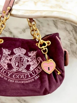 Lila Juicy Couture handväska i velour - Juicy Couture handväska i lila velour med broderad logga och emblem framtill. Väskan har guldfärgade kedjedetaljer, rosa läderremmar och ett hjärtformat hängsmycke med nyckel. Perfekt för dig som gillar en trendig och lyxig stil.
