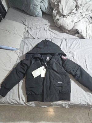 Svart dunjacka från Canada Goose - Säljer en svart dunjacka från Canada Goose med huva och ribbade muddar. Jackan har två stora fickor framtill, en ärmficka och den klassiska Canada Goose-loggan på ärmen. Perfekt för kalla vinterdagar och riktigt snygg streetstil.