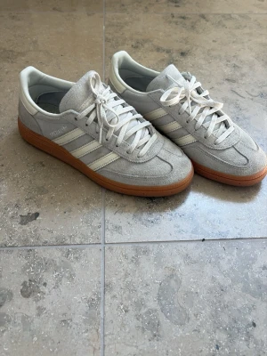 Adidas Spezial grå sneakers - Säljer ett par Adidas Spezial sneakers i ljusgrå mocka med vita detaljer och klassiska tre ränder på sidan. Skorna har vit snörning och en brun gummisula som ger en retrovibe. Perfekta för dig som gillar stilrena och tidlösa sneakers.