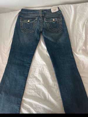 Blå bootcut jeans med kontrastsömmar - Säljer ett par blå bootcut jeans med tydliga kontrastsömmar och dekorativa fickor bak. Jeansen har en klassisk femficksmodell och är tillverkade i denim med lätt slitningar framtill och bak. Snygga detaljer på bakfickorna och normal midja.