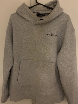 Grå hoodie från Sail Racing - Snygg grå hoodie från Sail Racing med broderad logga på bröstet. Klassisk känguruficka framtill och stor huva. Tillverkad i mjukt bomullsmaterial som är perfekt för chill dagar. Enkel och stilren design som passar till det mesta. Passar som S/M