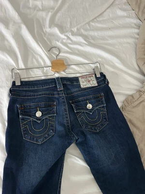 True Religion blå jeans med fickdetaljer - Säljer ett par blå jeans från True Religion med ikoniska kontrastsömmar och stora ficklock bak med silverfärgade knappar. Klassisk femficksmodell i denim med snygg tvätt och detaljerad brodyr på fickorna. Perfekta för en avslappnad och trendig look. säljer pga kommer inte till användning men super snygga jeans!💞