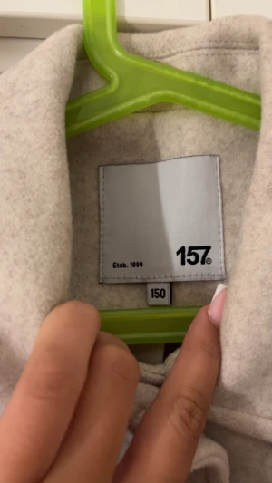 Beige ullkappa från 157 - Stilren beige ullkappa från 157 med klassisk krage och stora framfickor. Kappan har knäppning framtill och raka ärmar med knappdetalj vid ärmslut. Perfekt för kyligare dagar och enkel att matcha med olika outfits.