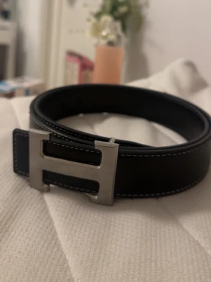 Svart skärp med silverfärgat H-spänne - Snyggt svart bälte med ett stort H-format spänne i silverfärg. Bältet har vita kontrastsömmar längs kanterna och är gjort i skinn. Perfekt accessoar för att lyfta din outfit.
