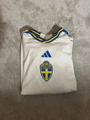 Svensk landslagströja Adidas vit - Vit svensk landslagströja från Adidas med blå och gula detaljer. Tröjan har korta ärmar, rund halsringning med blå/gula ränder och Sveriges landslagsemblem på bröstet. Perfekt för match eller träning.