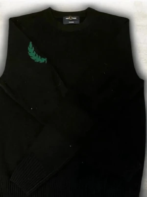 Fred Perry original - En svart stickad tröja med rund hals och ribbade muddar. Tröjan har en grön broderad lagerkrans på ena axeln som ger en unik touch. Perfekt för dig som gillar stilrena plagg med en liten twist.