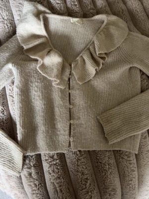 Beige kofta med volangkrage - Supersöt beige stickad kofta med volangkrage och knappar framtill. Koftan har långa ärmar och är gjord i ett mjukt, ribbat material som känns riktigt mysigt. Perfekt för dig som gillar en romantisk och trendig stil.