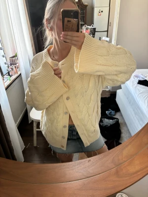 Oversize kabelstickad kofta i offwhite - Säljer en oversize kabelstickad kofta i ljus gul med stora knappar framtill. Koftan har rund halsringning, breda ärmar och ett mjukt, tjockt material som känns riktigt cozy. Perfekt att slänga över en t-shirt eller linne för en chill look.