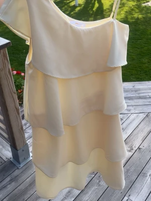 Ljusgul omlottklänning med volanger - Söt ljusgul klänning med volanger i flera lager och asymmetrisk axel, där ena sidan har smalt axelband och den andra är offshoulder. Klänningen har en luftig och flowy känsla, perfekt för varma dagar. Materialet ser ut att vara polyester. Liten fläck men den syns inte om man har utsläppthår. Pris kan diskuteras