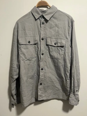 Grå overshirt från H&M - Säljer en stilren grå overshirt från H&M med två stora bröstfickor och knappar framtill. Skjortan har klassisk krage och långa ärmar med knapp vid ärmslut. Perfekt att bära öppen eller stängd för en avslappnad look.