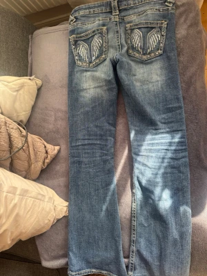 Blå jeans med broderade fickor - Säljer ett par blå jeans med raka ben och snygga slitningar. Baksidan har två fickor med broderade vingar i vitt och silver, vilket ger en unik och cool look. Jeansen har klassisk femficksmodell och kontrastsömmar. Perfekta för dig som vill sticka ut lite extra.