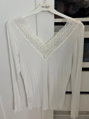 Vit långärmad topp med spets - Säljer en vit långärmad topp med v-ringning och bred spetskant runt halsen. Toppen är ribbad och har en feminin look som passar perfekt till jeans eller kjol. Materialet känns mjukt och stretchigt.