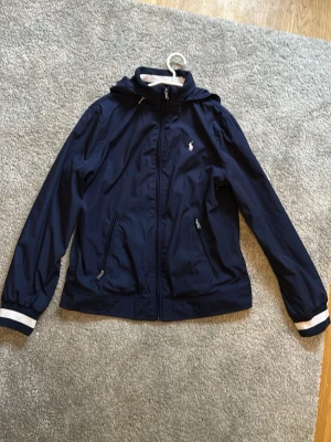 Marinblå vindjacka från Polo Ralph Lauren - Snygg marinblå vindjacka från Polo Ralph Lauren med vit logga på bröstet, huva och dragkedja framtill. Jackan har vita detaljer vid krage och ärmslut samt två fickor med dragkedja. Perfekt för blåsiga dagar.