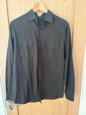 Mörkbrun skjorta med fickor - Stilren mörkbrun skjorta med två bröstfickor och knappar framtill. Skjortan har klassisk krage och långa ärmar, perfekt för en clean och enkel look. Tillverkad i ett mjukt material som känns bekvämt hela dagen.