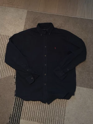 Mörkblå skjorta med knappar - Klassisk mörkblå skjorta med knappar framtill och lång ärm i storlek S. Skjortan har en liten broderad logga på bröstet och button down-krage. Perfekt till jeans eller chinos för en clean look.