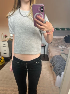 Grå cropped stickad t-shirt - Säljer en enkel och stilren grå stickad t-shirt med korta ärmar och rund hals. Tröjan är croppad och har en mjuk, lite luddig yta som ger en mysig känsla. Perfekt att matcha med höga jeans eller kjol.