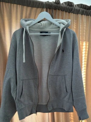 Vintage Ralph Lauren Hoodie - Snygg Vintage grå zip-up hoodie från Polo Ralph Lauren som har några år på nacken. Snygg vinröd logga.  Storlek M/M passar dock S. Mjuk och varm med fleecefodrad insida, huva, dragkedja och två framfickor. Ralph Lauren-loggan sitter på bröstet. Ett slitage vid ärmen men det stör inte.
