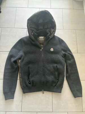 Moncler Tricot Cardigan - Snygg grå pufferjacka från Moncler med stickade ärmar och quiltad, glansig baksida och huva. Jackan har dragkedja framtill, två fickor med dragkedja och Moncler-logga på bröstet. Perfekt för kalla vinterdagar och riktigt trendig look. Köpt av Love Ekedahl på Facebook. Modellen heter Moncler G32-003 Padded Knit Down Jacket – Black/Grey – Size 1. Storlek L passar män strax över 180cm