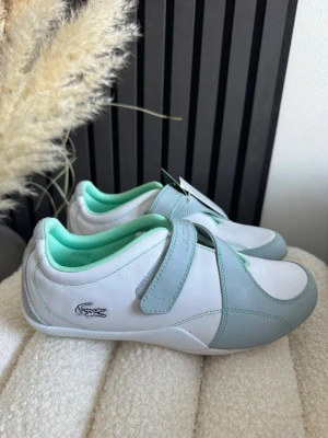 Vita och mintgröna Lacoste sneakers - Snygga sneakers från Lacoste i vitt och mintgrönt med kardborreband och klassisk krokodil-logga på sidan. Skorna har en låg profil och är tillverkade i slätt läder med mjukt textilfoder i mintgrönt. Perfekta för en clean och sportig look.