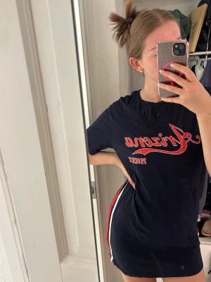 Marinblå kort t-shirtklänning - Säljer en marinblå kort t-shirtklänning med röd och vit text framtill och sportiga ränder längs sidorna. Klänningen har rund hals, korta ärmar och en avslappnad passform. Perfekt för en casual look.