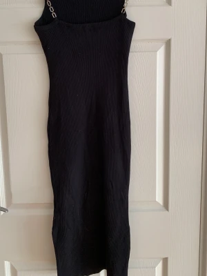 Svart ribbad klänning med kedjeband - Säljer en svart ribbad långklänning med smala axelband i kedjedesign. Klänningen är figurnära och har en stilren, minimalistisk look som passar perfekt till olika tillfällen. Materialet är stretchigt och bekvämt.
