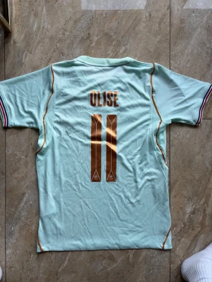 Frankrike fotbollströja Olise #11 Nike - Snygg ljusblå Frankrike fotbollströja med Olise och nummer 11 i guld på ryggen. Tröjan har korta ärmar, broderad tupp-logga och Nike-märke på bröstet samt tunna guldiga detaljer längs sidorna. Perfekt för match eller träning.