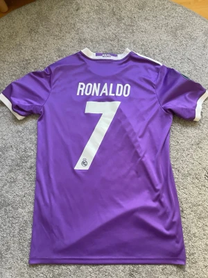 Lila Real Madrid Ronaldo matchtröja - Säljer en lila Real Madrid matchtröja från Adidas med Ronaldo och nummer 7 på ryggen. Tröjan har vita detaljer, klubbmärke, Champions League-patchar och Fly Emirates-tryck på bröstet. Tillverkad i funktionsmaterial som andas.