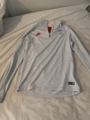 Vit långärmad Nike PSG träningströja - Snygg vit långärmad träningströja från Nike med PSG-logga på bröstet och röd Nike Swoosh. Tröjan har halv dragkedja framtill och är tillverkad i ett lätt, funktionsmaterial som passar perfekt till träning eller uppvärmning.