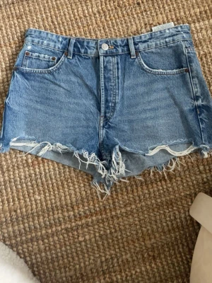 Blå jeansshorts med fransig kant - Säljer ett par klassiska blå jeansshorts med rå, fransig kant och femficksmodell. Shortsen har normal passform, bälteshällor och knappgylf. Perfekta för varma dagar och ger en avslappnad look.