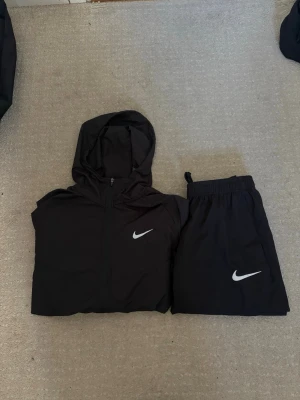 Svart Nike Miller set - Säljer ett svart träningsset från Nike med matchande jacka och byxor. Jackan har huva, dragkedja och två fickor, medan byxorna har resår i midjan och Nike-logga på benet. Perfekt för träning eller chill. Materialet är lätt och andas.