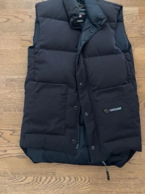 Svart dunväst från Canada Goose - Snygg svart dunväst från Canada Goose med två stora framfickor och diskret logga på vänster ficka. Västen har hög krage och stängs med både dragkedja och tryckknappar. Perfekt för lager-på-lager och håller dig varm under kyliga dagar.