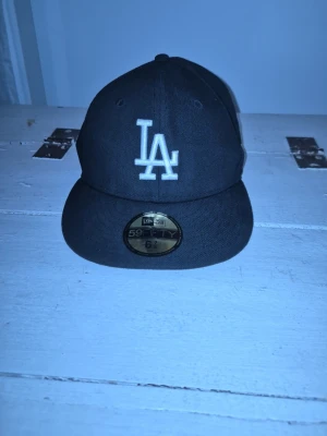 Svart LA keps 59FIFTY - Säljer en svart New Era 59FIFTY keps med LA-logga broderad i vitt framtill och MLB-logga bak. Klassisk flat peak och strukturerad passform. Perfekt för dig som gillar streetwear och sportig stil.