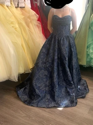 SÖKES! Mörkblå långklänning med blommönster  - Under 3000 kr. Elegant mörkblå långklänning med vackert blommönster och markerad midja. Klänningen har en hjärtformad urringning och är ärmlös med bred kjol som ger en klassisk siluett. Perfekt för bal eller festliga tillfällen.