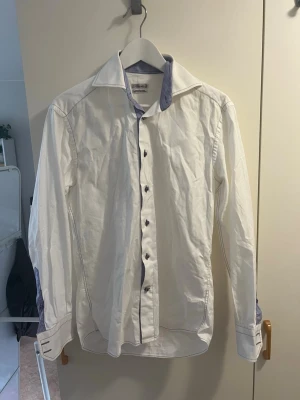 Vit skjorta med blå detaljer - Klassisk vit skjorta med blå rutiga detaljer på insidan av kragen och manschetterna. Skjortan har långa ärmar, knappar framtill och en snygg kontrast mellan vitt och blått. Perfekt för en stilren look.