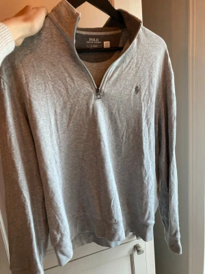 halvzip Ralph Lauren - Snygg grå långärmad tröja från Polo Ralph Lauren med halv dragkedja och klassisk broderad logga på bröstet. Tillverkad i mjukt bomullsmaterial och har en avslappnad passform. Perfekt till jeans eller chinos. 