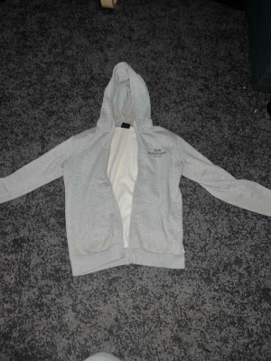 Ljusgrå zip hoodie med huva - Säljer en ljusgrå hoodie med dragkedja och huva. Tröjan har fickor framtill och texten 'Peak Performance' broderad på bröstet. Tillverkad i mjukt sweatshirtmaterial, perfekt för chill dagar. Enkel och stilren design som passar till det mesta.