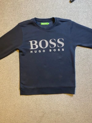 Mörkblå sweatshirt från Hugo Boss - Mörkblå sweatshirt med rund hals och stort BOSS-tryck på bröstet. Klassisk passform och ribbade muddar vid ärmslut och nederkant. Tillverkad i mjukt material för en bekväm känsla. Perfekt för en avslappnad och stilren look.