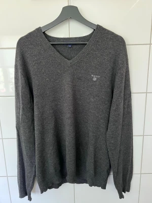 Mörkgrå stickad tröja från GANT - Mörkgrå stickad tröja från GANT med V-ringning och diskret logga broderad på bröstet. Tröjan har långa ärmar och ribbade muddar vid ärmslut och nederkant. Perfekt för lager på lager och en stilren look.
