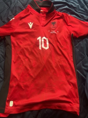 Albaniens matchtröja nr 10 röd - Säljer en röd matchtröja från Albaniens landslag med nummer 10 och namnet N. Bajrami på ryggen. Tröjan har vita detaljer, svart krage och logga på bröstet. Tillverkad i funktionsmaterial, perfekt för fotboll eller träning. Han använde den i en match men tyvärr har bokstäverna gått bort däremot är allt annat där 