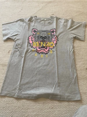Grå Kenzo t-shirt med tigertryck - Snygg grå t-shirt från Kenzo med ikoniskt tigertryck i svart, rosa och gult framtill samt texten 'KENZO'. Klassisk passform och rund hals. Perfekt för dig som vill sticka ut med en trendig och ungdomlig look. Det står XL men sitter som en S.