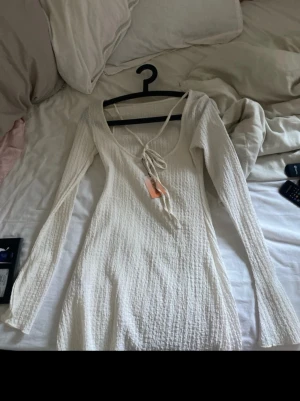 Vit långärmad ribbad klänning - Snygg vit kortklänning med lång ärm och ribbad struktur. Klänningen har en djup v-ringning med snörning framtill och är tillverkad i ett mjukt, stretchigt material som formar sig efter kroppen. Perfekt för dig som gillar en enkel men trendig look.