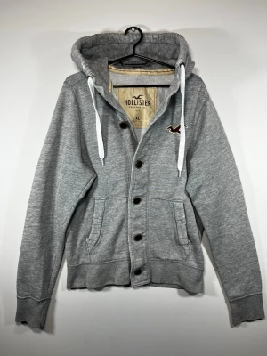 Hollister grå hoodie med knappar – XL - Grå Hollister hoodie i storlek XL med knappstängning, broderad Hollister-logga på bröstet och vit mjuk insida. Klassisk äldre Hollister-design med snygg vintage/Y2K-look.  Begagnat skick med tydliga bruksspår. Mindre hål/slitage på armbågarna, samt lite slitage på muddarna, se bilder. Fortfarande fullt användbar och slitaget passar bra till den vintage-inspirerade looken.  Storlek: XL, men skulle säga att den passar mer som L Färg: Grå