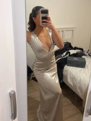 Champagnefärgad långklänning i satin - Säljer en elegant långklänning i champagnefärgad satin med djup v-ringning och ärmlös design. Klänningen har en figurnära passform och ett lyxigt, glansigt material som ger en exklusiv känsla. Perfekt för dig som vill sticka ut på nästa bal eller fest.