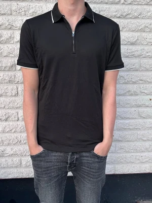 Svart polotopp med dragkedja - Snygg svart polotopp med kort ärm och dragkedja framtill. Kragen och ärmsluten har vita detaljer som ger en clean kontrast. Perfekt till jeans eller shorts för en enkel men stilren look. Storlek S. 