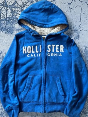 Blå hoodie från Hollister med huva - Blå zip-hoodie från Hollister med vit broderad text på bröstet och mjukt teddyfoder i huvan. Två fickor framtill och dragkedja hela vägen. Väldigt skön!💕Inga skador eller något sånt endast att fluffet är inte lika fluffigt som de va när den köptes!
