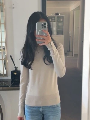 Beige stickad tröja med knappar - Snygg beige stickad tröja med långa ärmar och dekorativa knappar vid ärmsluten. Tröjan har en smal passform och rund halsringning, perfekt att matcha med jeans eller kjol.