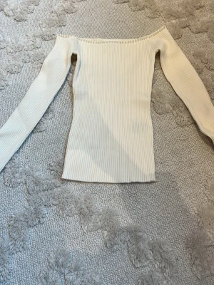 Vit ribbad offshoulder topp - Trendig vit ribbad offshoulder topp med lång ärm. Sitter tight och har en snygg, figurnära passform. Perfekt att styla med jeans eller kjol för en clean look.
