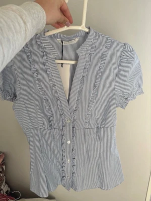 Blårandig blus med volang från Zara - Söt blårandig blus från Zara med korta puffärmar och volangdetaljer längs knäppningen. Blusen har smal passform, rund halsringning och tunna vertikala ränder i vitt och blått. Perfekt till jeans eller kjol för en fräsch look. Blusen är enbart testad och är ny med prislapp
