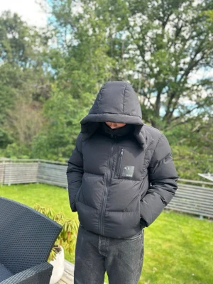 Svart pufferjacka från Armani Exchange - Säljer en svart pufferjacka från Armani Exchange med stor huva och dragkedja framtill. Jackan har en bröstficka med logga, två sidofickor och en boxig passform. Perfekt för kalla dagar och riktigt snygg streetstil. Justerbar midja och luvan kan man ta bort ifall man vill