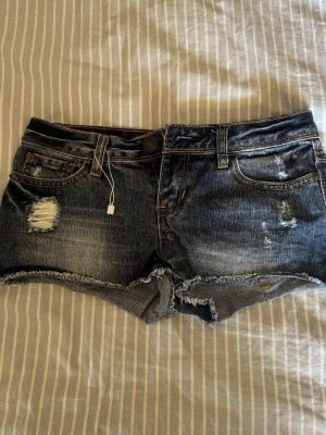 Blå slitna jeansshorts Aeropostale - Säljer ett par blå jeansshorts från Aeropostale med slitna detaljer och råa kanter. Shortsen har fem fickor, knapp och dragkedja framtill samt klassisk jeanslook med kontrastsömmar. Perfekta för varma dagar och en avslappnad stil.
